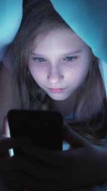 Dikey video. Çocuk bağımlılığı. Çevrimiçi gece. Odaklanmış, takıntılı bir kız. Akıllı telefonuyla battaniyenin altına saklanıyor. Evde yatarken içeriği izliyor..