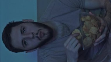 Dikey video. Gece filmi. Amatör. Rahat adam televizyon seyrediyor. Mavi ışıklı, karanlık bir odada abur cubur yiyor..