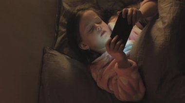 Dikey video. Yatma vakti. Gece aleti. Uykusuz çocuk. Yorgun küçük kız, gece geç saatte yatakta telefonda çizgi film izliyor..