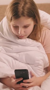 Dikey video. Çocuk bağımlılığı. Ev tatili. Dijital yaşam tarzı. Ciddi kız sabahları yatakta battaniyenin altında akıllı telefondan besleniyor..