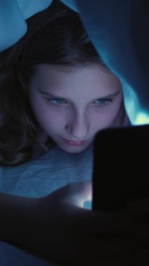 Dikey video. Ekran zamanı. Çocuk uykusuzluğu. Geç saate kadar. İnternet bağımlılığı. Uykusuz, yorgun kız akıllı telefondan mesajlaşıyor evde battaniyenin altında saklanıyor..