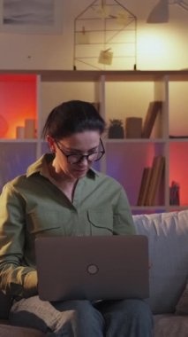 Dikey video. İnternet kullanımı. Ağ taraması. Genç kız açıklayan kadın dizüstü bilgisayar kullanıyor. Akşam kanepede oturuyor..