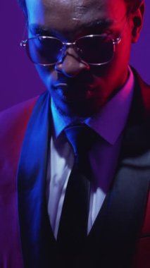 Dikey video. Neon light adam klas tarzı. Harika görünüyor. Zarif kırmızı smokinli özgüvenli beyefendinin portresi koyu floresan mor gözlüklü..