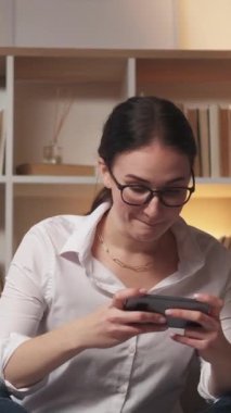 Dikey video. Gezici oyun. Ev tatili. Gadget hafta sonu. Heyecanlı, hayal kırıklığına uğramış bir kadın modern iç mekandaki koltukta oynarken telefonunu kaybediyor..