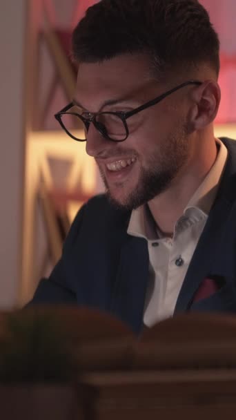 垂直ビデオ オンライン通信 笑顔の男 ホームワーク ハッピースマート男タイプラップトップ座ってデスクでオープン本で暗い部屋のインテリア — ストック動画