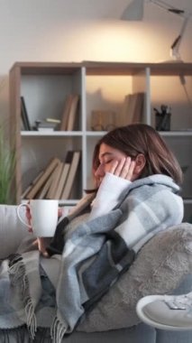 Dikey video. İğrenç bir çay. Virüs içiyor. Boğaz ağrısı. Titreyen kadın soğuk algınlığına yakalanıyor elinde bardak tutarak iyileştirici grip evinde kanepeye sarılı olarak oturuyor..