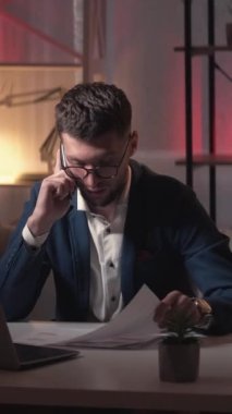 Dikey video. Mobil iletişim. Çalışan adam. Bilgileri kontrol et Akıllı iş adamı dokümanları inceliyor. Akıllı telefon konuşuyor. Karanlık ışık odasının iç tarafında oturuyor..