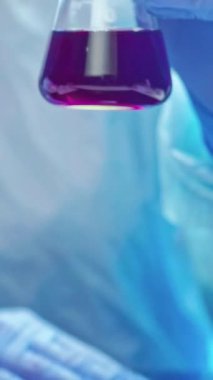 Dikey video. Laboratuvar araştırması. Uyuşturucu geliştirme. Tıbbi kimya. Koruyucu eldivenli teknisyen ellerindeki dolu cam şişedeki tepkiyi inceliyor..