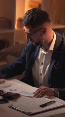 Dikey video. Muhasebe süreci. Erkek bir uzman. Finansal hesaplamalar. Yoğunlaşmış adam notlar alıyor. Belgeler üzerinde çalışıyor. Karanlık ışık odasının iç tarafında oturuyor..