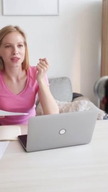 Dikey video. Blog öğretmenliği. Dil hocası. Masada notlar ve dizüstü bilgisayarla gülümseyen kendine güvenen bir kadın, kamera içeriğiyle konuşuyor..