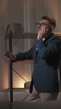 Dikey video. Hoş bir iletişim. Mutlu adam. Evde rahatla. Gülümseyen adam rahat görünmeli. Karanlık odada akıllı telefondan konuşmalı..