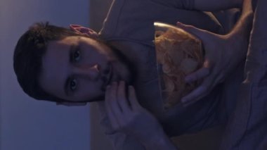 Dikey video. Gece filmi. Ev tatili. Meraklı ve endişeli bir adam, TV şovunu izlerken gecenin geç saatlerinde, tek başına, mavi ışıklı karanlıkta abur cubur yiyor..
