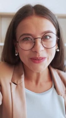 Dikey video. Vlog içeriği. Etkileyici akış. Neşeli bir şekilde gülümseyen, neşeli bir genç kadın. Gözlükleriyle kameraya el sallıyor..