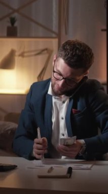 Dikey video. İş görevleri. Meşgul adam. Finansal muhasebe. Zengin adam cep telefonuyla konuşan nakit para sayıyor. Karanlık odanın içinde oturuyor..