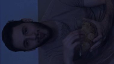 Dikey video. Film gecesi. Ev eğlencesi. Keyifli adam, komik komedi dizilerini izlerken eğleniyor. Mavi ışıklı karanlık odada cipsleri yerken gülüyor..