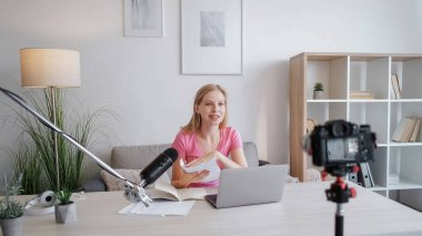 Koçluk blogu. İçerik kaydı. Masadaki kendine güvenen genç kadın notlarıyla tripodda kameraya ve mikrofona konuşan ışık odasında video çeken..