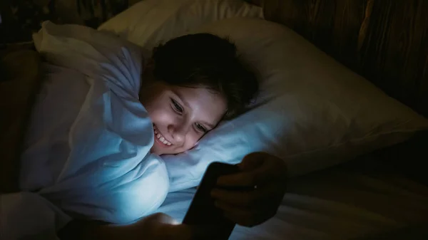 子供中毒だ デジタルレジャー 夜のインターネットサーフィン 幸せな笑顔女の子スクロールソーシャルメディアフィード上のスマートフォン嘘でベッドに暗い部屋に自宅 — ストック写真