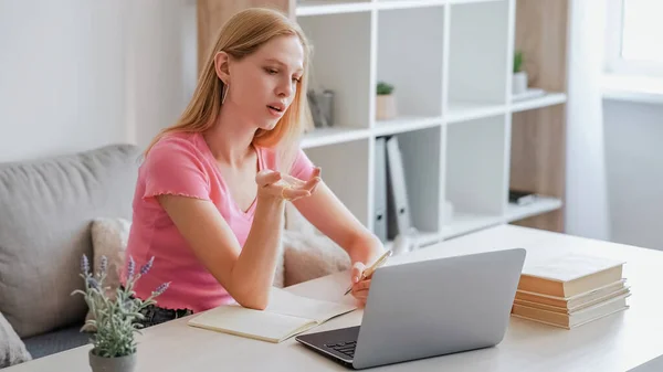 インターネット研究 学ぶことに挑戦 自己教育 軽いインテリアでノートパソコンやノートブックとテーブルで手に質問ジェスチャーとペンで座っている若い女の子を混乱 — ストック写真