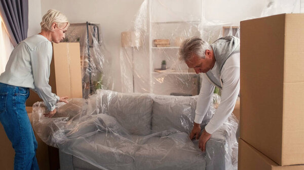 Переезжаю. Снимаю дом. Муж супружеской пары, покрывающий диван полиэтиленовой пленкой упаковочные принадлежности светлый интерьер комнаты.