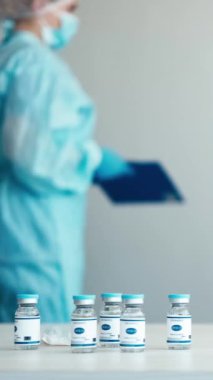 Dikey video. Covid-19 aşısı. Klinik politikası. Salgını durdurun. İlaç şişeleri beyaz masada. Mavi üniformalı hemşire ve maske takan kadın..