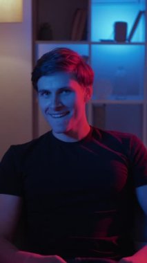 Dikey video. Harika bir plan. Üçkağıtçı adam. Meme ifadesi. İğneleyici bir adam ellerini ovuşturup şakayı düşünüyor. Kanepede oturuyor. Koyu neon ışıklı ev yapımı..