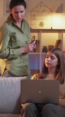 Dikey video. Bilgisayar açıklıyor. Laptop yardımı. Genç kızı gösteren bir kadın dizüstü bilgisayar programı kullanıyor. İnternette akşam kanepede oturuyor..