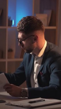 Dikey video. İş için. Erkek başhekim. Bilgi çalış. Zeki sıradan adam notlar alıyor. Belgeleri kontrol ediyor. Karanlık ışık odasının iç tarafında oturuyor..