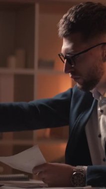 Dikey video. Bilgileri. Endişeli adam. İş başarısız oldu. Gerekli belgeleri bulmak için kendini çaresiz hisseden meşgul bir adam karanlık odanın içinde masa başında oturuyor..