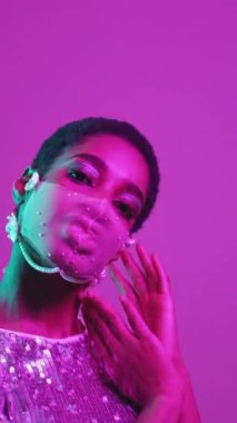 Dikey video. Pandemik moda. Karantina aksesuarı. Şenlikli güzellik. Mor pembe neon ışıklı cazibeli kadın. Ağlı maske takıyor. Parlak pullu elbise..