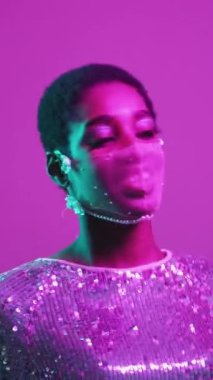 Dikey video. Salgın partisi. Disko görüntüsü. Karantina eğlencesi. Mor pembe renkli neon ışıklı cazibe neon maskeli parlak elbiseyle dans eden mutlu kadını eğlendirdi..