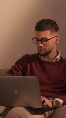 Dikey video. Mesafe çalışması. Zeki, sıradan bir adam. Bilgisayar teknolojisi. Yakışıklı adam dizüstü bilgisayarda çalışıyor. Koltuğun gölgesinde oturuyor..
