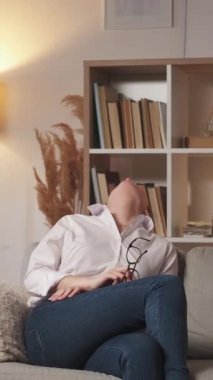 Dikey video. Yorgun dinlenme. Ev baş ağrısı. İyi akşamlar yorgunluğu. Yorgun öğretmen kadın başını okşayarak modern oturma odasındaki boş alana sahip kanepeye oturuyor..