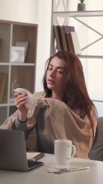 Dikey video. Enfeksiyon kapmış. Soğuk kadını yakalamak. Çevrimiçi toplantı. Hasta kadın, masa başında oturmuş hapşırıyor ve çay içiyor. İç mekanda dizüstü bilgisayarla konuşuyor..