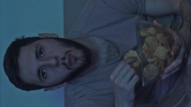 Dikey video. Gece filmi. Amatör. Rahat adam televizyon seyrediyor. Mavi ışıklı, karanlık bir odada abur cubur yiyor..