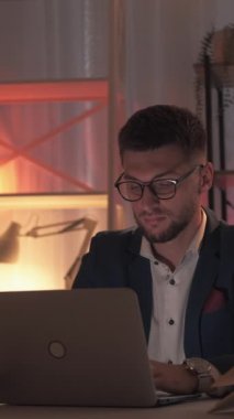 Dikey video. Mesafe çalışması. Meşgul adam. Araştırma bilgileri. Masanın üzerinde karanlık odanın içinde açık kitaplarla dizüstü bilgisayarı yazan konsantre bir adam..