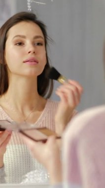 Dikey video. Güzellik makyajı. Sabah rutini. Gülümseyen genç bir kadın aynaya dekoratif makyaj fırçasıyla kontur tozu sürüyor..