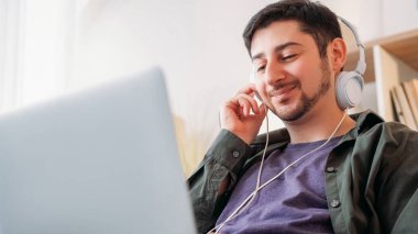 Müzik aleti. Şarkı keyfi. Cihaz eğlencesi. Neşeli gülümseyen kaygısız adam evdeki modern laptopta kulaklıkla mp3 müzik listesi dinliyor..