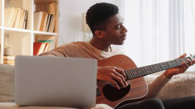 Çevrimiçi müzik okulu. Gitar öğretmeni. Uzaktan öğrenme. Dizüstü bilgisayarda telli enstrüman dersi veren adam evdeki kanepede video görüşmesi yapıyor..