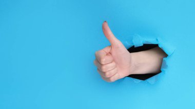 Jest gibi. Olumlu geri bildirim. Mavi yırtık kağıt deliği arka planında onaylı başparmak gösteren kadın eli boş alan.