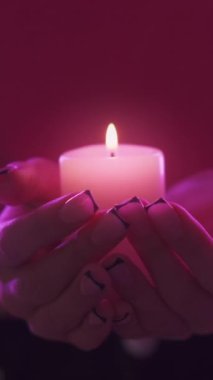 Dikey video. Mum meditasyonu. Dua geleneği. Kadın ellerinin parlak pembe neon ışıkta yanan mum ışığını tuttuğu yakın plan.