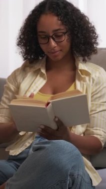 Dikey video. Okuma hobisi. Rahat bir ortam. Evde rahatla. Neşeli kadın, oturma odasında en sevdiği kitapla ev huzurunun keyfini çıkarıyor..