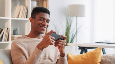 Gezici oyun. Gadget eğlencesi. Cihaz eğlencesi. Neşeli adam online esport uygulaması oynuyor. Akıllı telefondan ev oturma odasındaki kanepede program izliyor..