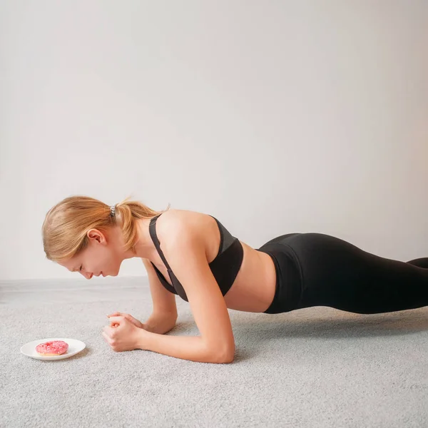 排気食 スポーツの動機 減量の訓練 疲れた女が床の上で板を持って苦労し 甘いドーナツデザートは空のスペースにあります — ストック写真