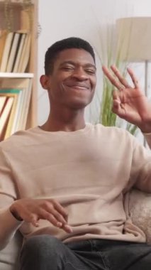 Dikey video. Onaylandı. Tamam jest. Memnuniyet verici genç adam evdeki oturma odasındaki kanepede tavsiye yazısı gösteriyor..