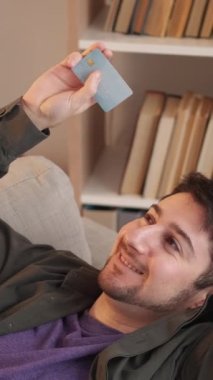 Dikey video. Kredi kartı servisi. Banka müşterisi. Plastik hesap numarası. Keyifli bir adam kanepede dinleniyor. İnternetten nakit geri ödemeli ev kredisi almaktan memnun..