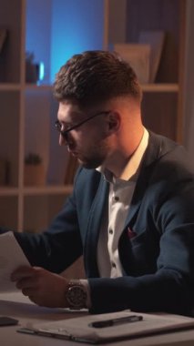 Dikey video. İş için. Erkek başhekim. Bilgi çalış. Zeki sıradan adam notlar alıyor. Belgeleri kontrol ediyor. Karanlık ışık odasının iç tarafında oturuyor..