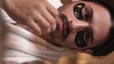 Dikey video. Göz bantları. Yüz bakımı. Yaşlanma karşıtı cilt bakımı. Sakallı, kendine güvenen, bıyıklı, nemlendirici siyah hidrojel pedleri süren, cildine dokunan bir adam..
