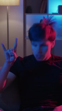 Dikey video. Boynuz hareketi. Rock 'çı adam. Meme ifadesi. Komik asık suratlı adam, punk müzikten zevk alıyor. Kanepe neon ışıklı odada oturuyor. İçi döngü dolu..