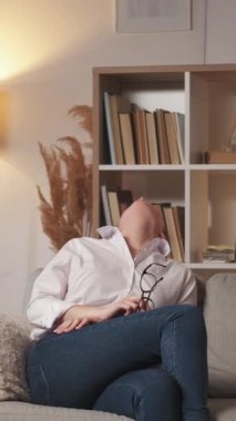 Dikey video. Yorgun dinlenme. Ev baş ağrısı. İyi akşamlar yorgunluğu. Yorgun öğretmen kadın başını okşayarak modern oturma odasındaki boş alana sahip kanepeye oturuyor..