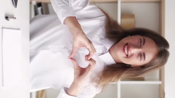 垂直ビデオ 愛を送る オフィスの女性 インスピレーションを得た作品 笑顔女性でシャツに座ってデスクショーハート形の手で光の部屋のインテリア — ストック動画